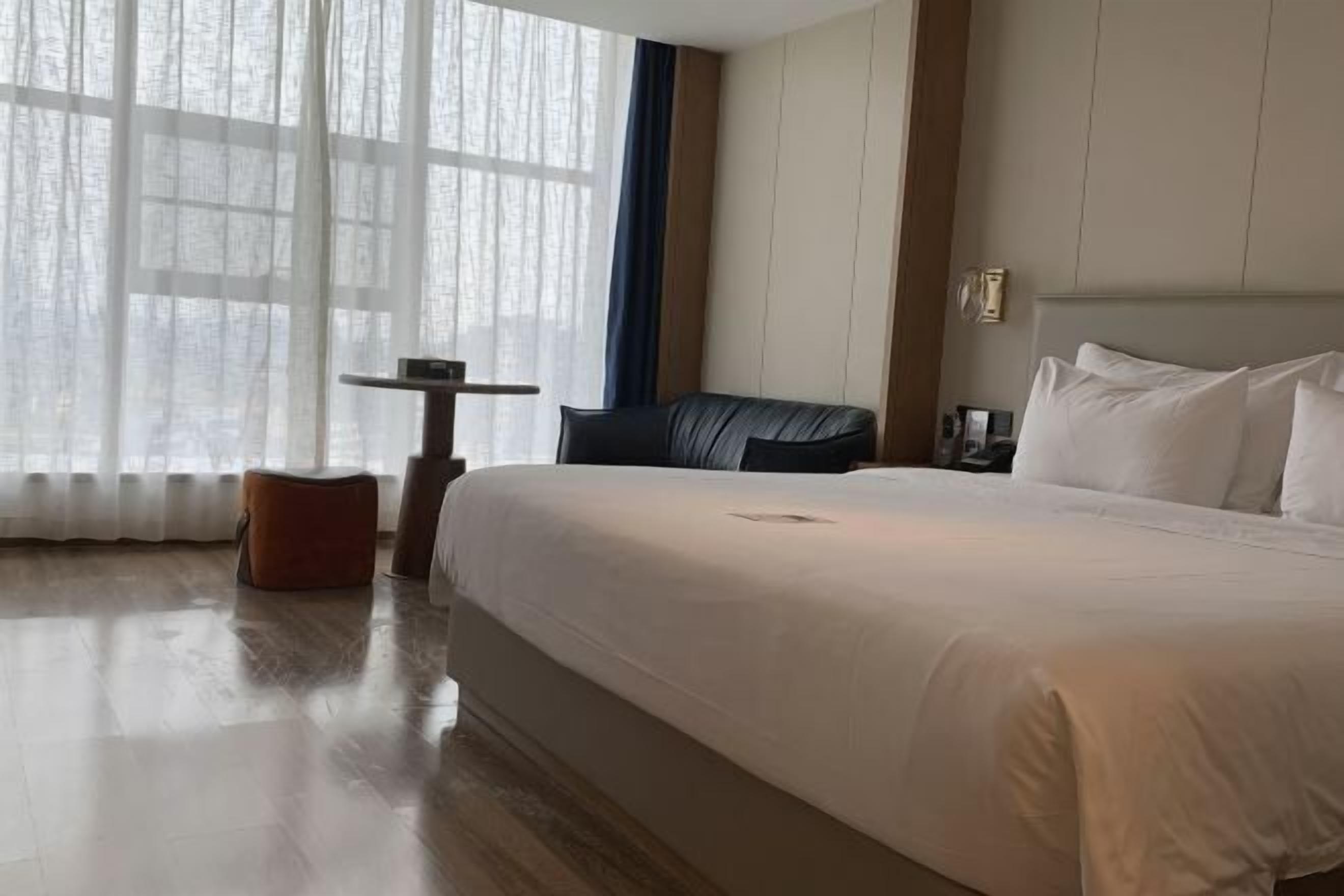 Una habitación de hotel con una cama grande, una mesita pequeña, una silla y una ventana con cortinas translúcidas.