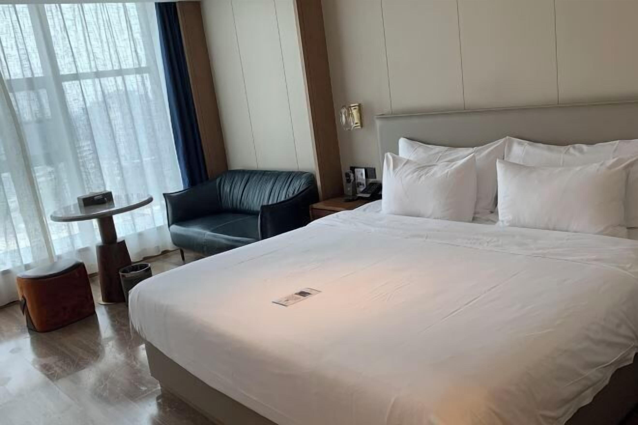 A hotel room with a large bed, a small table, a chair, and a window with curtains.