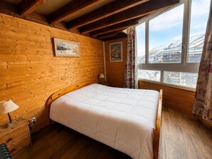 A wooden-paneled room with a bed, a nightstand, a window with a view of a snowy landscape, and two framed pictures on the wall.