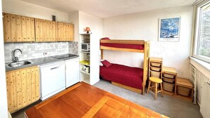 A compact kitchen with wooden cabinets, a sink, and a microwave. A bunk bed with red bedding is in the corner, and a small table with a chair is next to it.