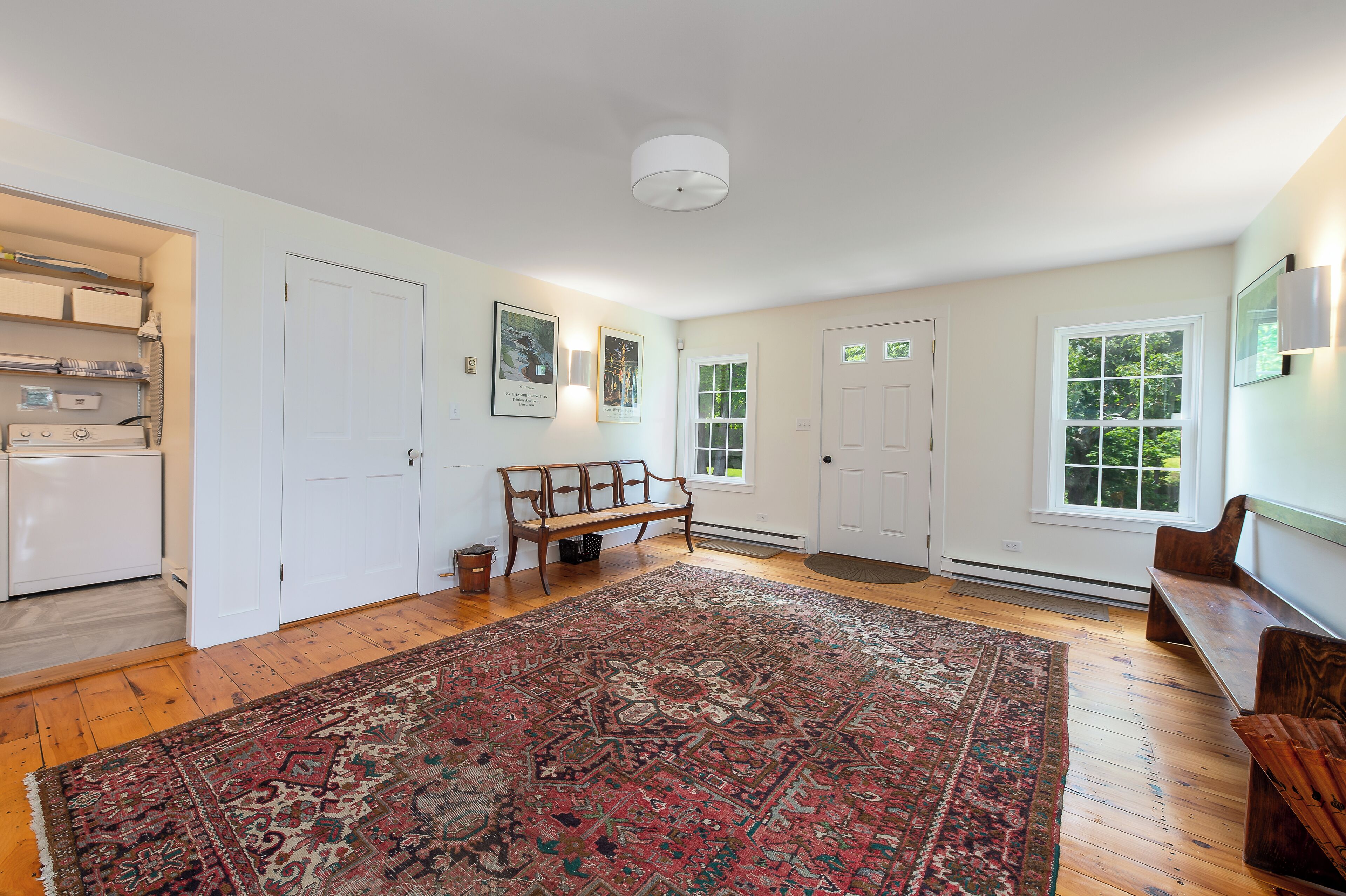 A spacious room with hardwood flooring, a large patterned rug, a bench, and a view of a garden through a window.