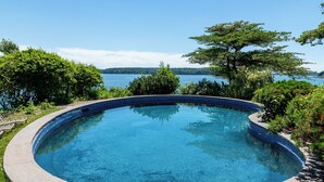 Une piscine circulaire à eau d’un bleu limpide, entourée d’une végétation luxuriante et offrant une vue sur un lac.