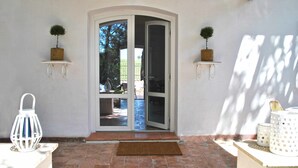 A doorway with a glass door leading to a patio area. There are two small potted plants on either side of the door, a white lantern on the left, and a white table with a basket on the right. - VILLA CORBEZZOLO between nature and the sea (Sperlonga)