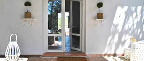 A doorway with a glass door leading to a patio area. There are two small potted plants on either side of the door, a white lantern on the left, and a white table with a basket on the right.