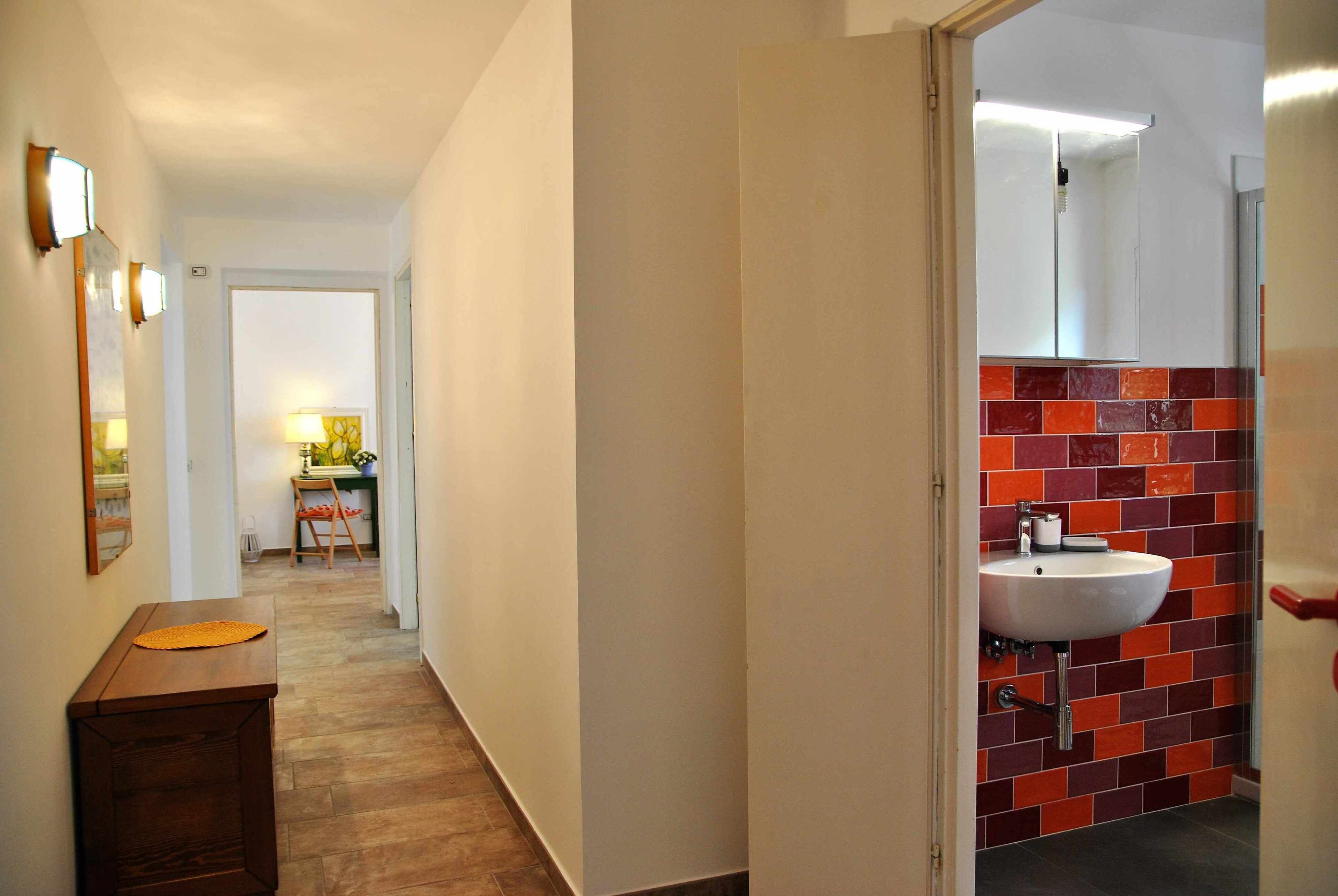 Un pasillo con un banco de madera, un lavabo blanco y un salpicadero de azulejos rojos, y un espejo encima.