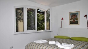 A bedroom with a bed, green pillows, a window with a view of trees, and a painting on the wall. - VILLA CORBEZZOLO between nature and the sea (Sperlonga)