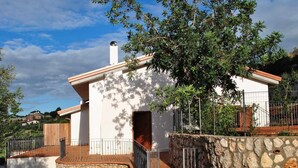 A small, white building with a stone wall and metal railing, surrounded by trees and a clear sky.