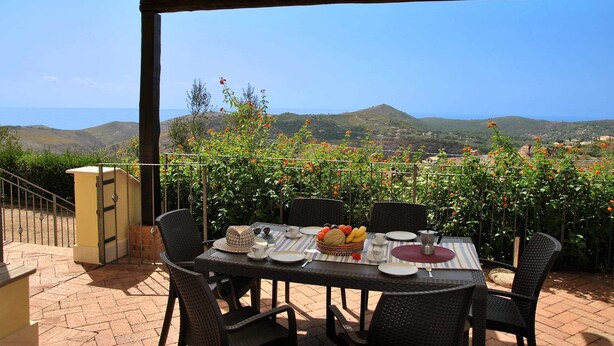 An outdoor patio with a table set for dining, surrounded by chairs, and overlooking a scenic view of hills and the sea.