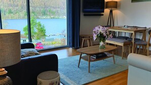 A living room with a large window overlooking a lake, a flat-screen TV mounted on the wall, a wooden coffee table, and a sofa. - 14 Loch View in Kenmore (Aberfeldy)
