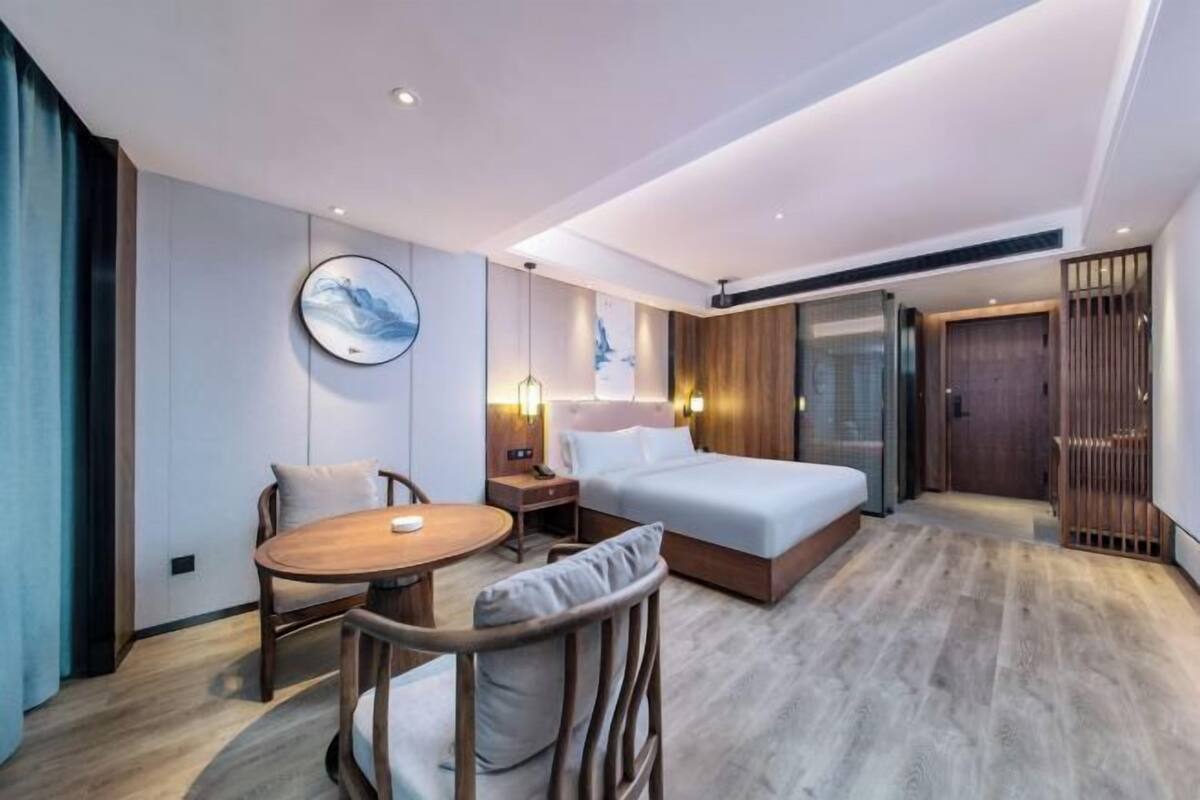 A modern hotel room with a bed, a round dining table, a chair, a nightstand, and a wall-mounted artwork.