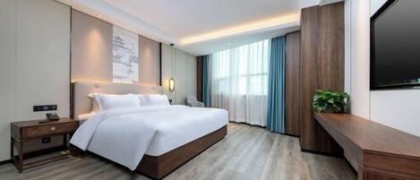 Habitación de hotel moderna con una cama grande, una mesita de noche con lámpara, un televisor de pantalla plana en la pared y un ventanal con cortinas.