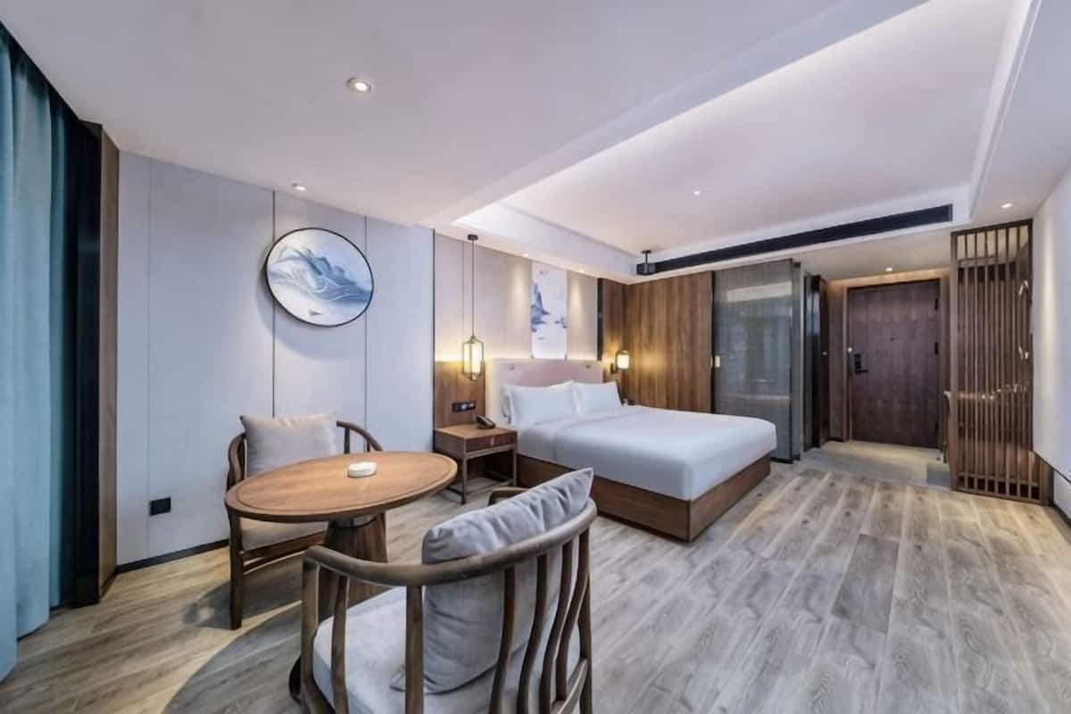 A hotel room with a bed, a round wooden table, a chair, a nightstand, and a wall-mounted lamp.