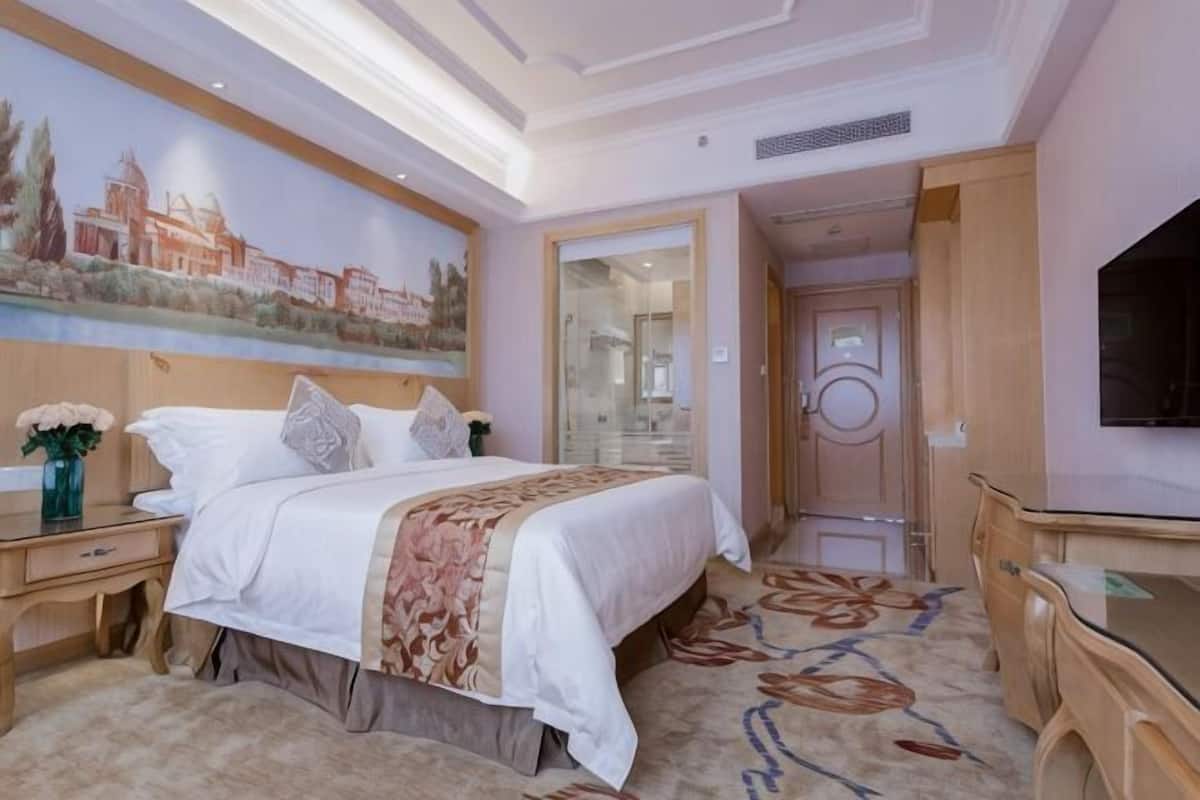 A hotel room with a bed, a nightstand, a TV, a desk, and a painting on the wall.