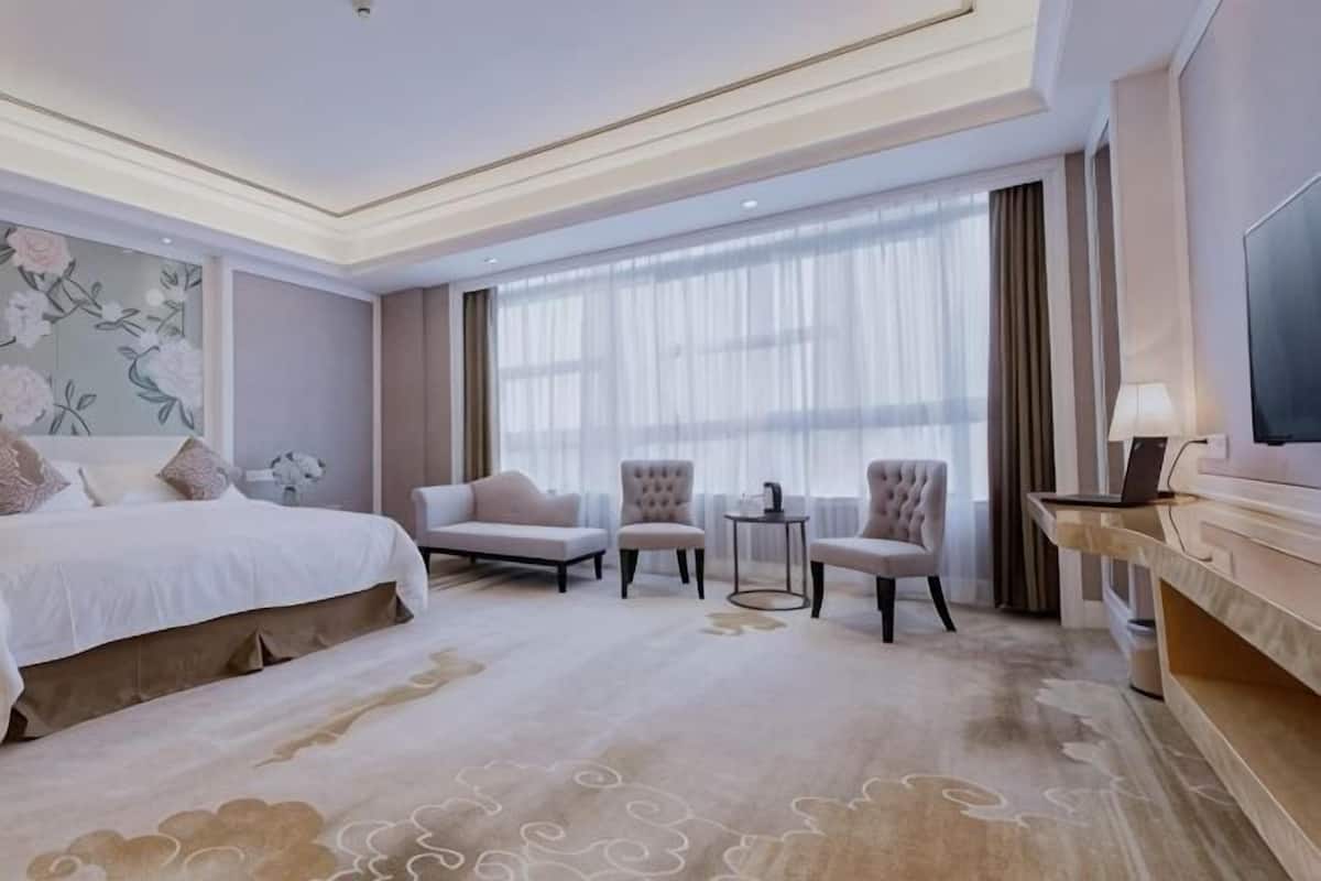 A hotel room with a large bed, two armchairs, a small table, a TV, and a large window with curtains.