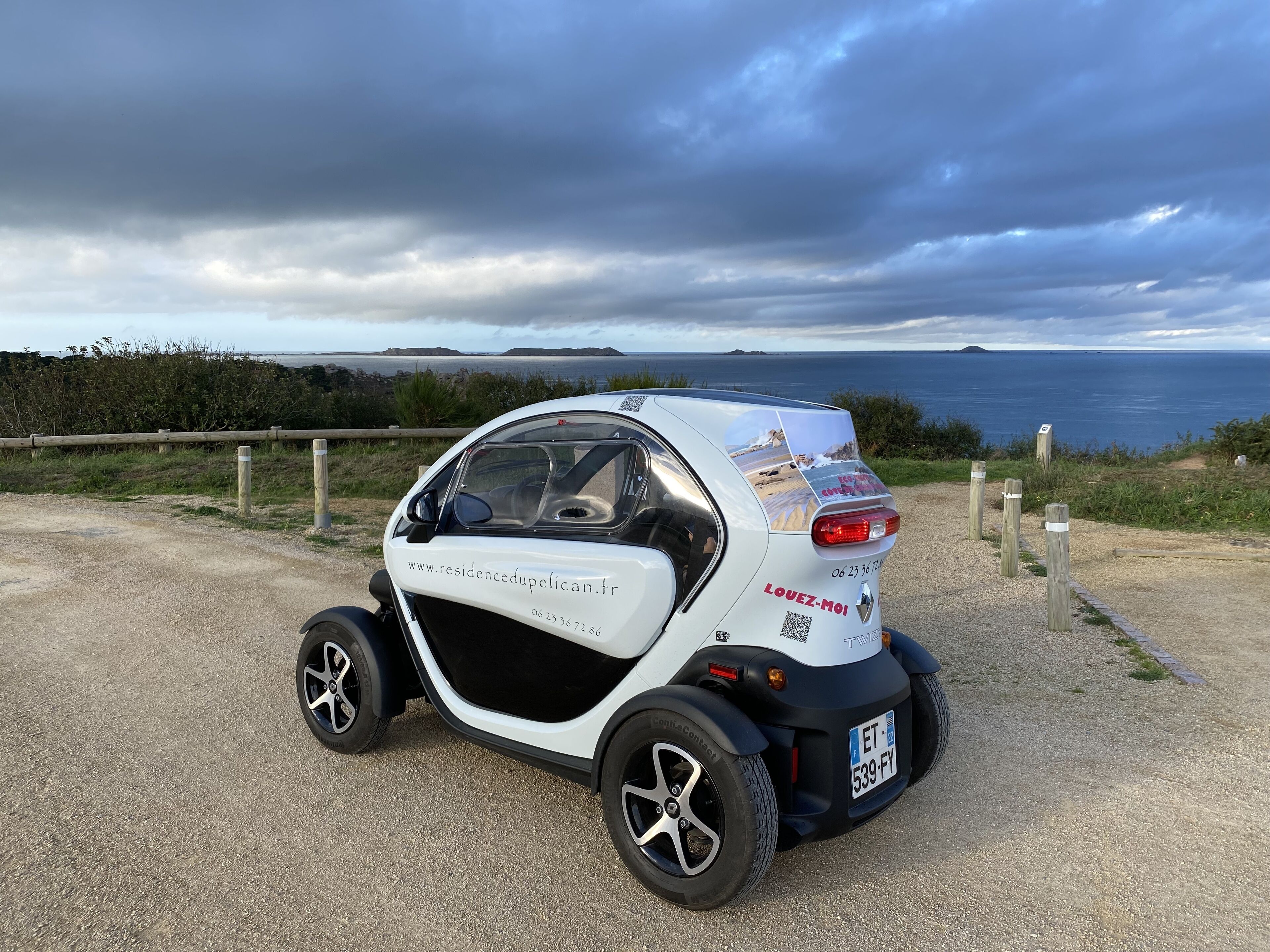 Een kleine, witte auto geparkeerd op een grindpad, met een prachtig uitzicht op de kust op de achtergrond.
