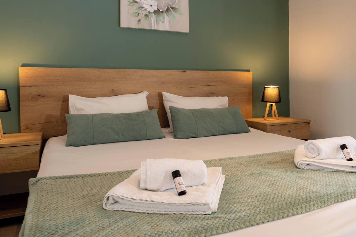 Una cama bien hecha con dos almohadas, una manta verde y dos toallas dobladas, con una botella en cada lado.