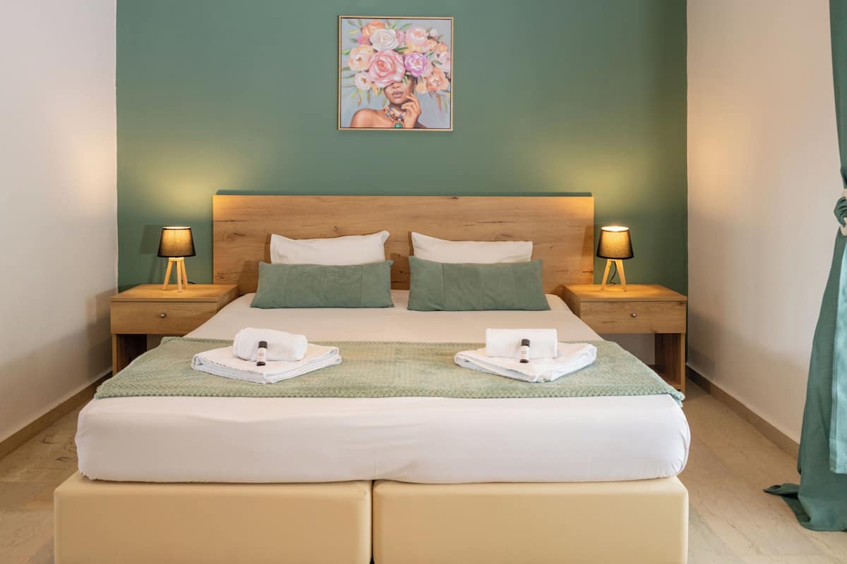 Una cama doble con sábanas bien puestas, dos almohadas, una mesita de noche con lámpara y un cuadro en la pared.