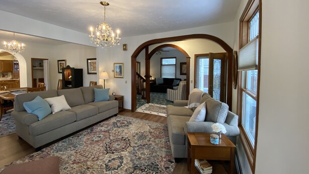 A spacious living room with a large sofa, a smaller sofa, a coffee table, and a patterned rug. There is a chandelier hanging from the ceiling, and a dining area is visible in the background. - Betsy Ross,, Great Downtown,, Residence (Gaylord)