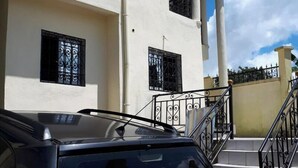 A black SUV parked in front of a two-story building with a white facade, black window grills, and a staircase leading to the entrance. - Comfortable duplex / 160 m2 (Douala)
