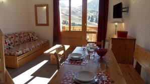 A cozy room with a wooden sofa, a dining table set with plates, glasses, and cutlery, and a view of mountains through a large window. - 100m from ski slopes, 4th floor, view ski slopes, balcony, tv, ski locker, 32m², La Plagne (La Plagne)