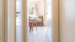 Un couloir mène à une pièce dont le sol est recouvert de carreaux, et qui contient un canapé, une table et une chaise.