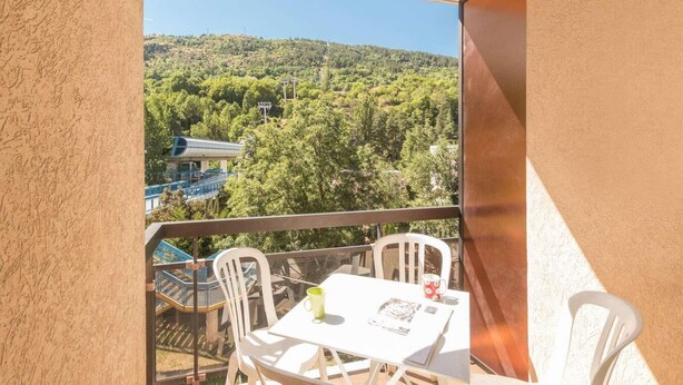 Un balcon avec une table et des chaises, donnant sur une zone boisée et un bâtiment avec un toit bleu.