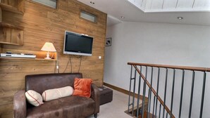 A living room with a brown leather sofa, a flat-screen TV mounted on a wooden wall, a lamp, and a wooden staircase with a metal railing.