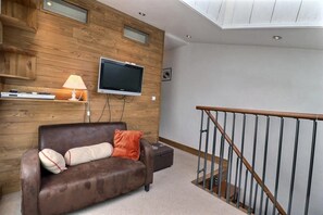 A living room with a brown leather sofa, a flat-screen TV mounted on a wooden wall, and a wooden staircase with a metal railing. - 10m from ski slopes, 4th floor, balcony, 55m², Méribel-Mottaret (Méribel - Les Allues)