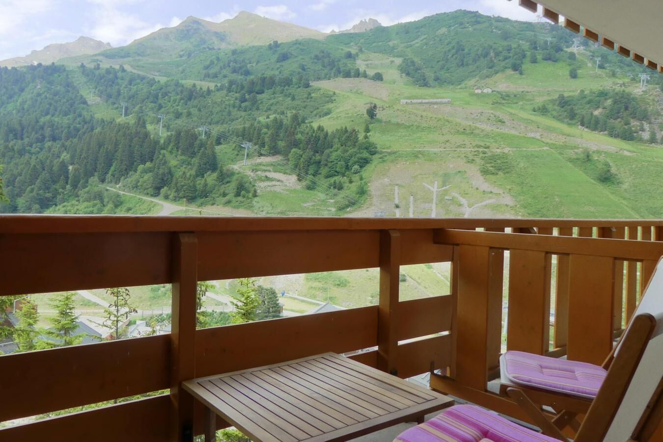 A balcony with a wooden table and a cushioned chair, overlooking a mountainous landscape with greenery and a road.