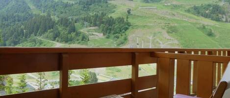 A balcony with a wooden table and a cushioned chair, overlooking a mountainous landscape with greenery and a road.