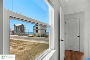 A room with a large window overlooking a beach and buildings, a white door, and a wooden floor.