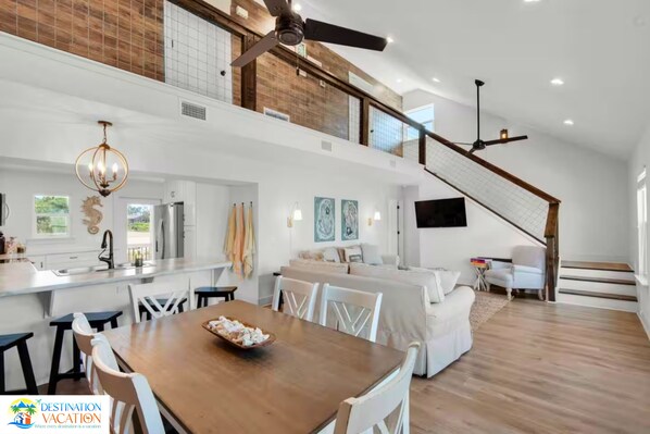 A spacious living area with a dining table, white furniture, and a staircase leading to an upper level.