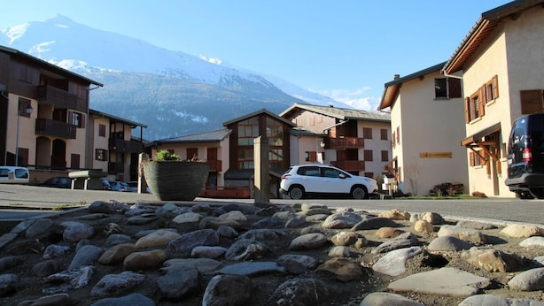 A mountain village with traditional buildings, a parked car, and a large rock garden in the foreground.
