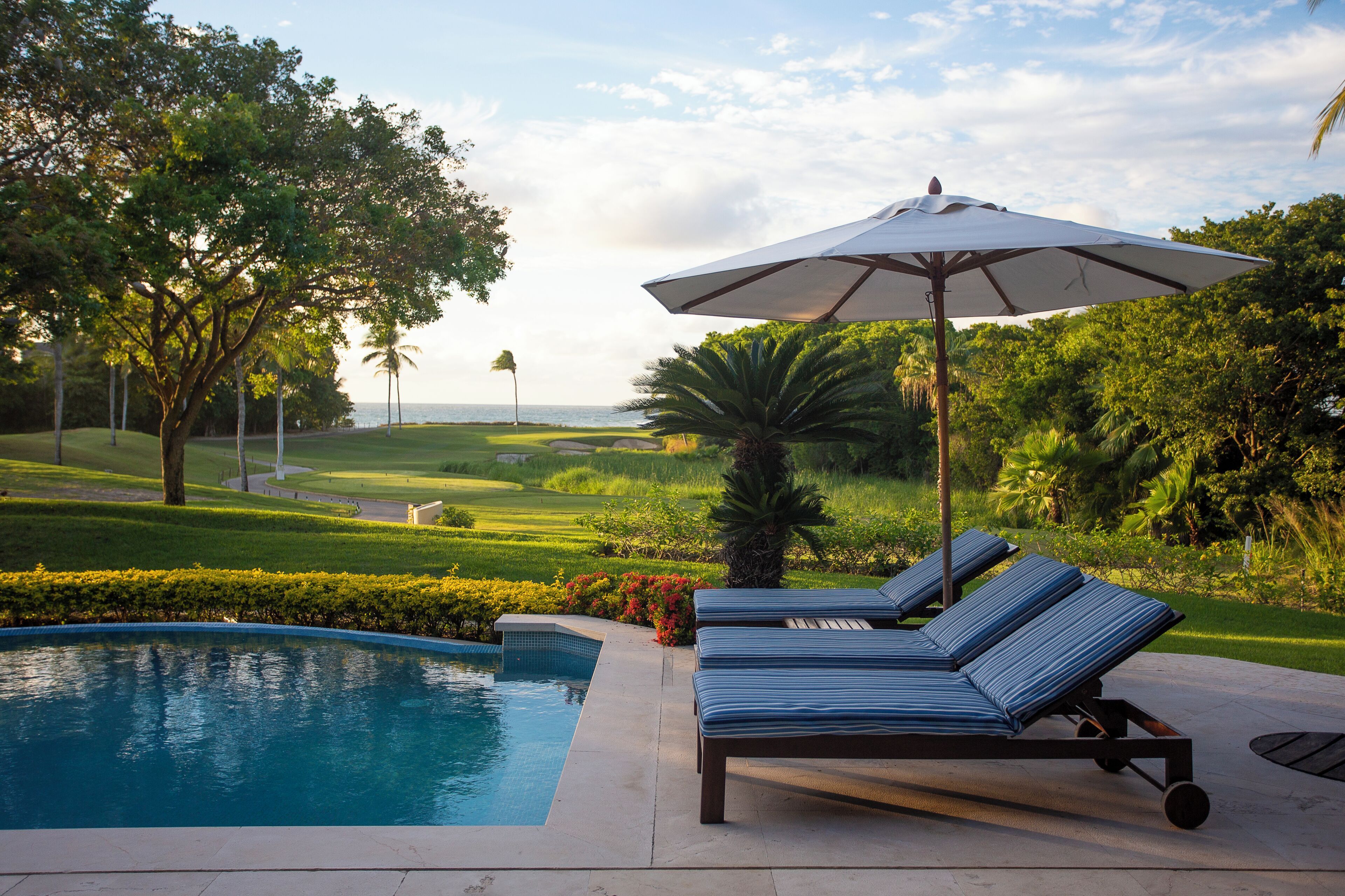 Uma área à beira da piscina com espreguiçadeiras e um guarda-sol, cercada por vegetação e vista para o oceano.