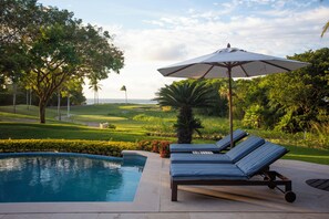 A poolside area with lounge chairs and an umbrella, surrounded by greenery and a view of the ocean.