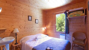 A cozy wooden cabin bedroom with a bed, a bedside lamp, a window with a view of greenery, a shelf with folded clothes, and a wicker chair. - Chalet, 250m from ski lifts, sauna, terrace, balcony, fireplace or stove, tv, 130m², Les 2 Alpes (Vénosc)
