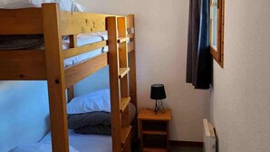 Une chambre avec un lit superposé, une petite table et une lampe, une fenêtre avec des rideaux et un radiateur.