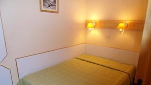 Ein kleines Zimmer mit einem Einzelbett, einer grünen Tagesdecke, zwei Wandleuchten und einem gerahmten Bild an der Wand.