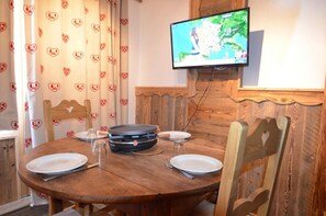 A dining area with a round wooden table, four chairs, a television mounted on the wall, and heart-patterned curtains.