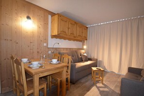 A cozy room with a wooden dining table set for four, a sofa, and a small wooden stool. - 10m from ski slopes, 8th floor, view ski slopes, balcony, tv, 24m², Les Menuires (Les Menuires)