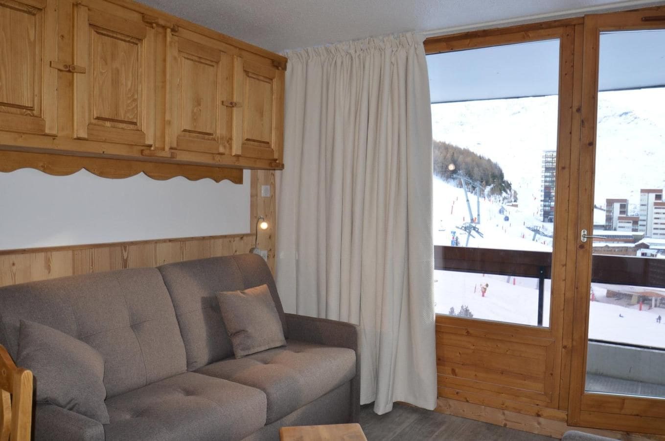 A wooden cabin interior with a sofa, curtains, and a view of a snowy landscape.