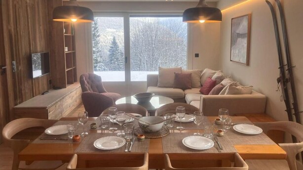 Un comedor con una mesa redonda dispuesta para seis personas, rodeada de sillas de madera. Se observa un sofá y una televisión al fondo.