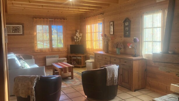 Interior acogedor de una cabaña de troncos, con un sofá, dos sillones, una mesita, un televisor y una zona de cocina.