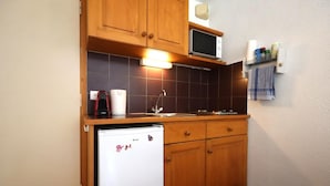 A compact kitchen with wooden cabinets, a microwave, a refrigerator, and a small shelf with kitchen items.