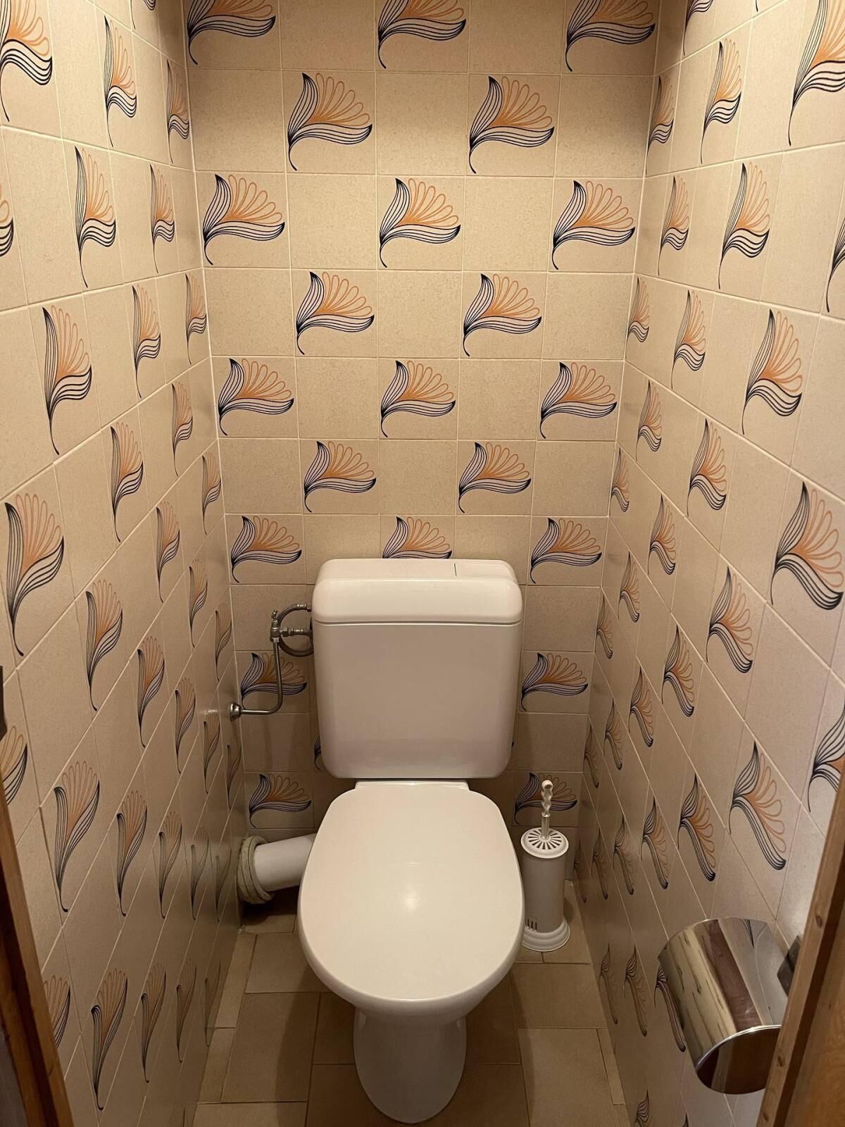 A small bathroom with a toilet, a bidet, and a trash can. The walls are tiled with a floral pattern.