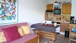 Une pièce chaleureuse avec un canapé en bois, un banc, une table à manger recouverte d’une nappe noire et un coin cuisine doté de meubles en bois et d’un four à micro-ondes.