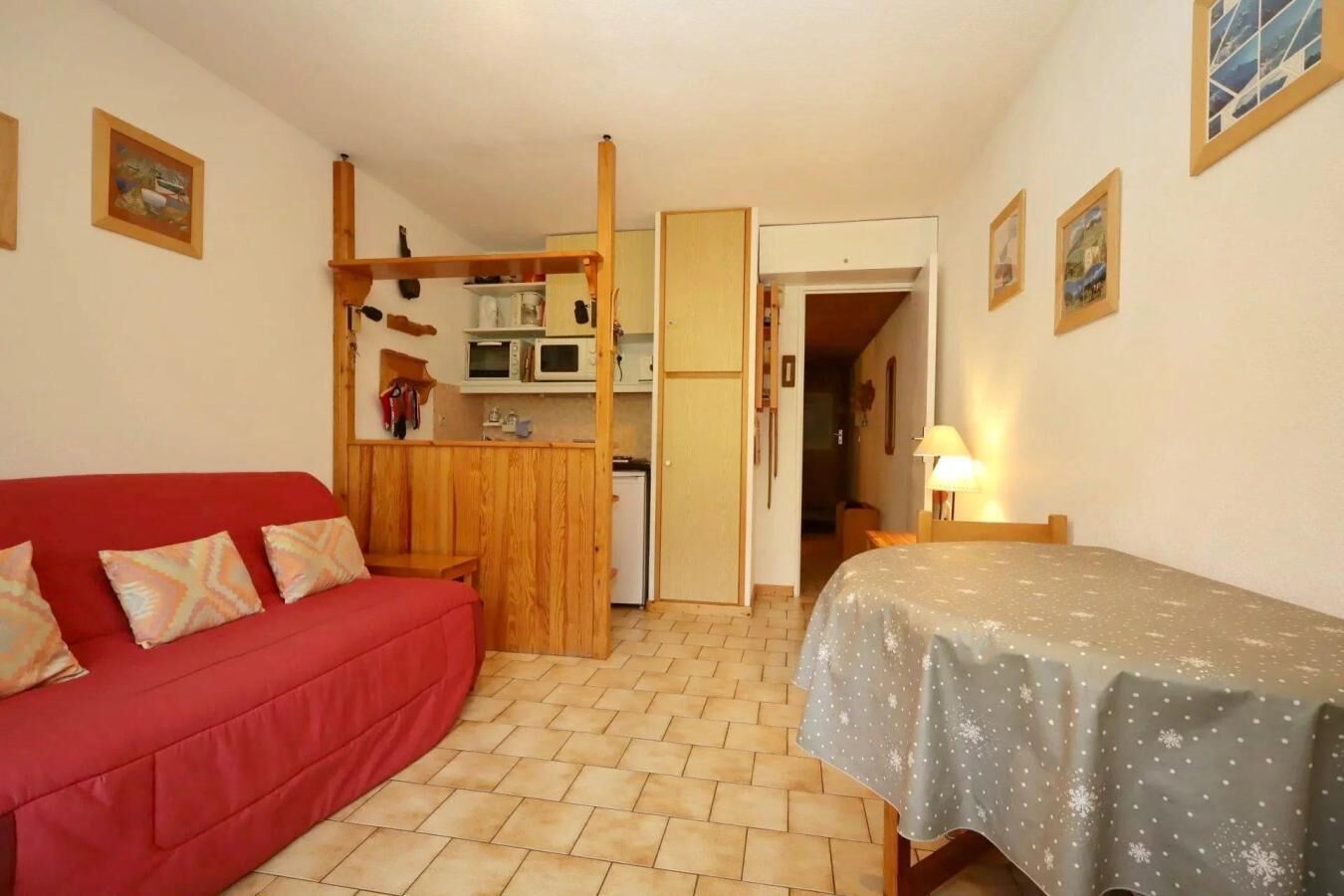 Een compacte kamer met een rode bank, een bed met een sterrenbedekking, een kitchenette met een magnetron, en een houten deur die naar een andere kamer leidt.