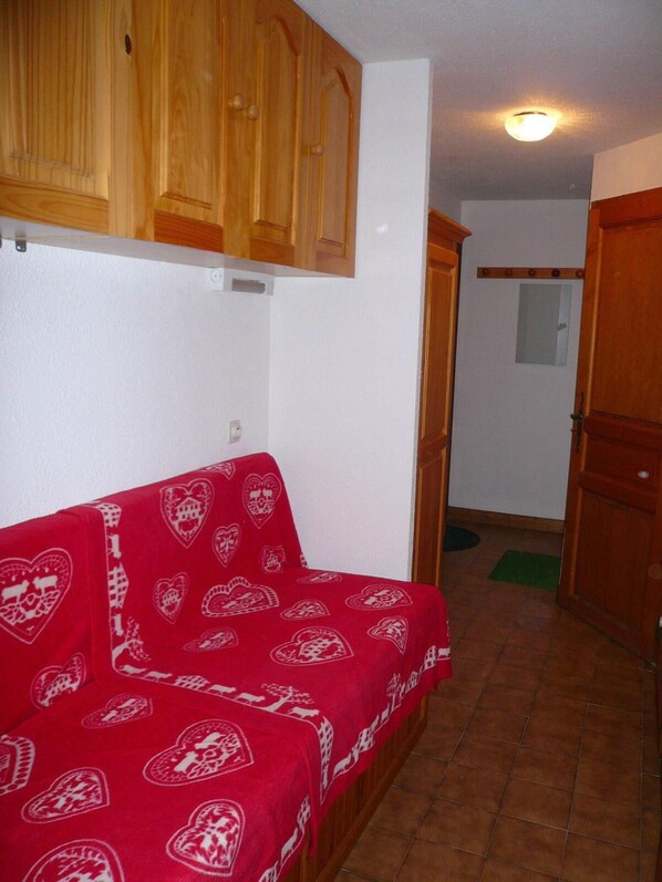A room with a red sofa covered in a patterned blanket, wooden cabinets, a tiled floor, and a door leading to another room.