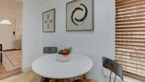 Un comedor con una mesa redonda, dos obras de arte enmarcadas, una planta en maceta y una ventana con persianas.
