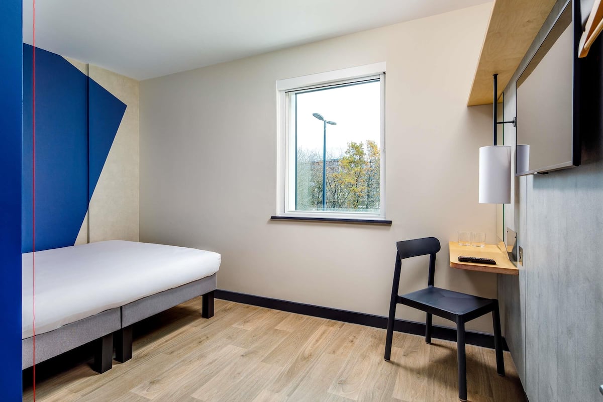 A small, modern room with a bed, a desk, and a chair. There is a window providing natural light and a view of trees outside.